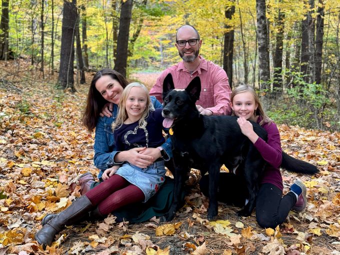 Nate Thames with his wife, their two daughters and their black dog, sitting in fallen leaves in the woods.